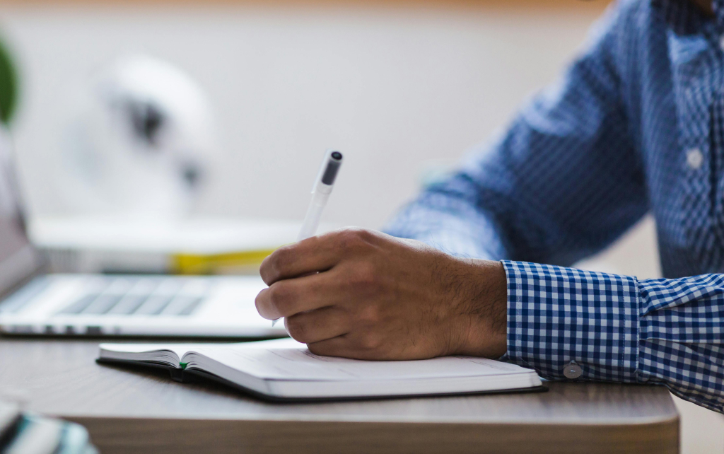 A man writing in a notebook with a pen next to an open laptop.