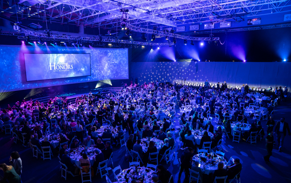 Overview of the 2025 President's Honors event, showing guests in a room at circular tables facing a stage where a screen shows the event logo