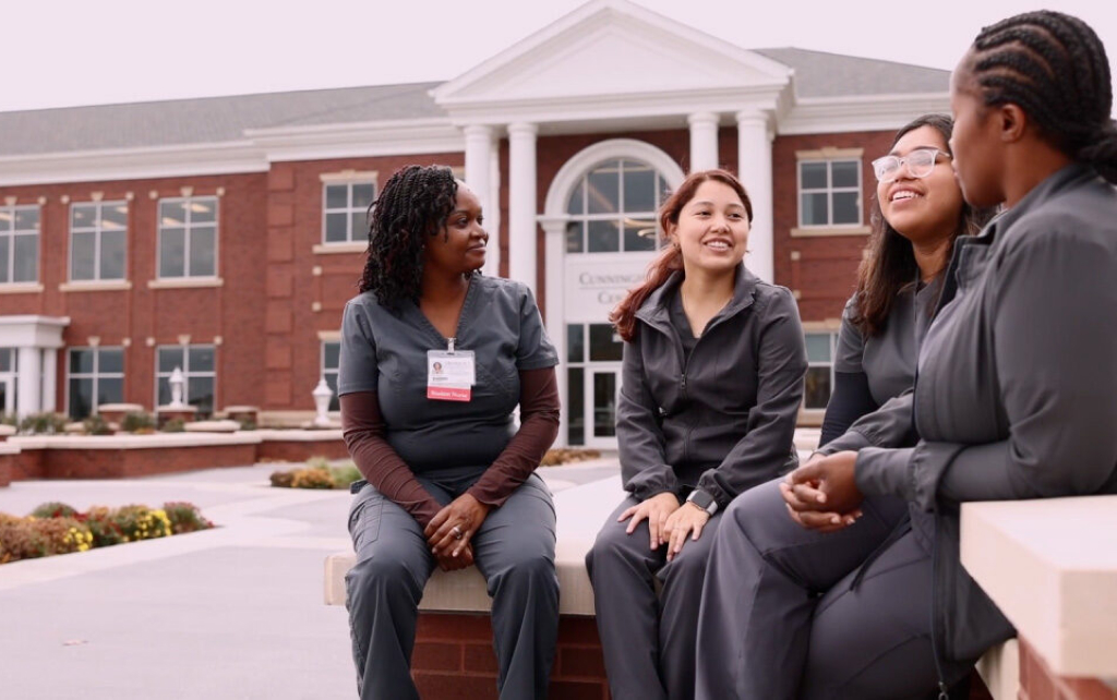 A group of nursing students in gray scrubs sit on a brick wall having a conversation out in front of the Cunningham Center on the MNU campus