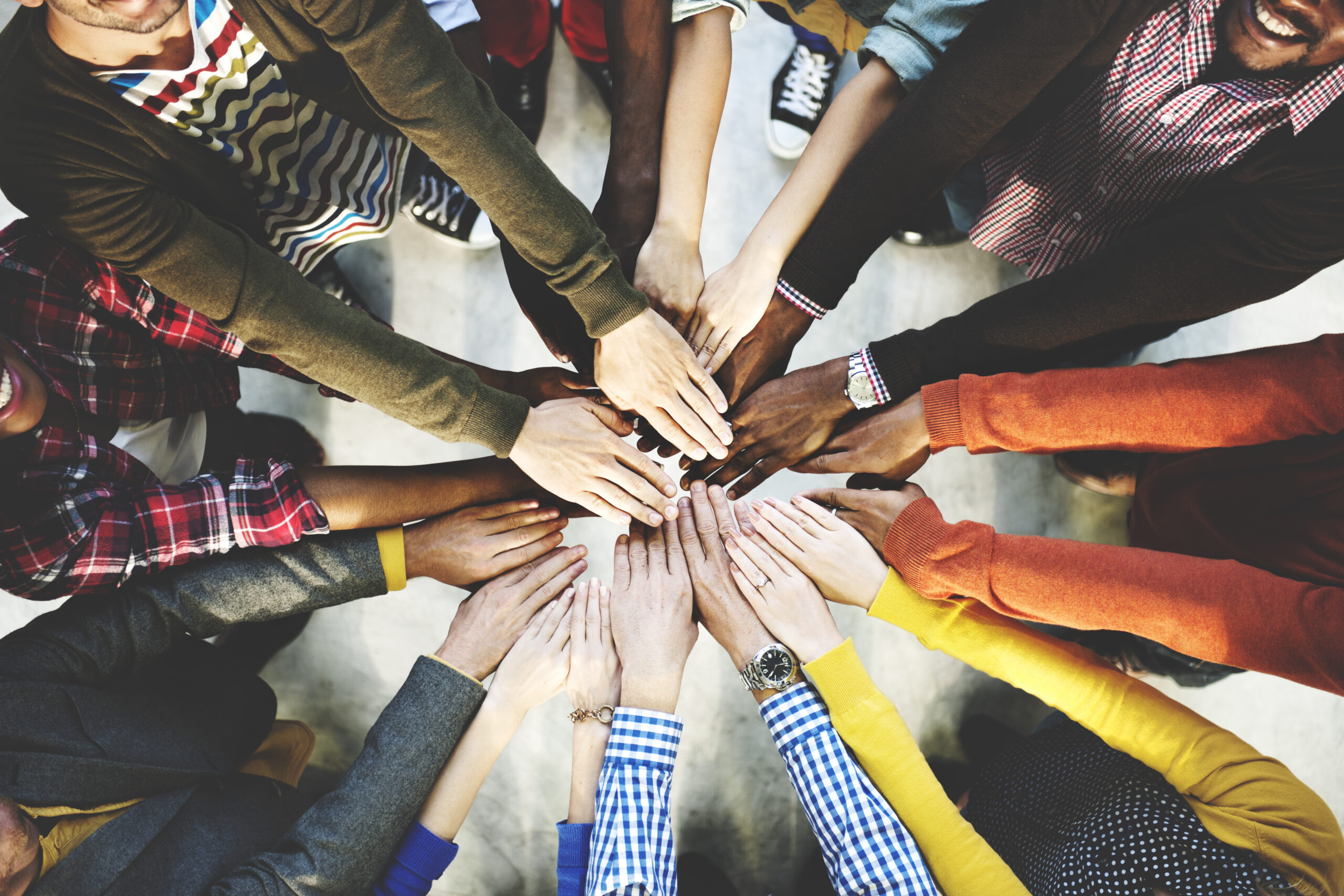 A group of people stand together in a circle with their hands outstretched into the middle. The group is diverse, composed of different skin types and people wearing colorful shirts.